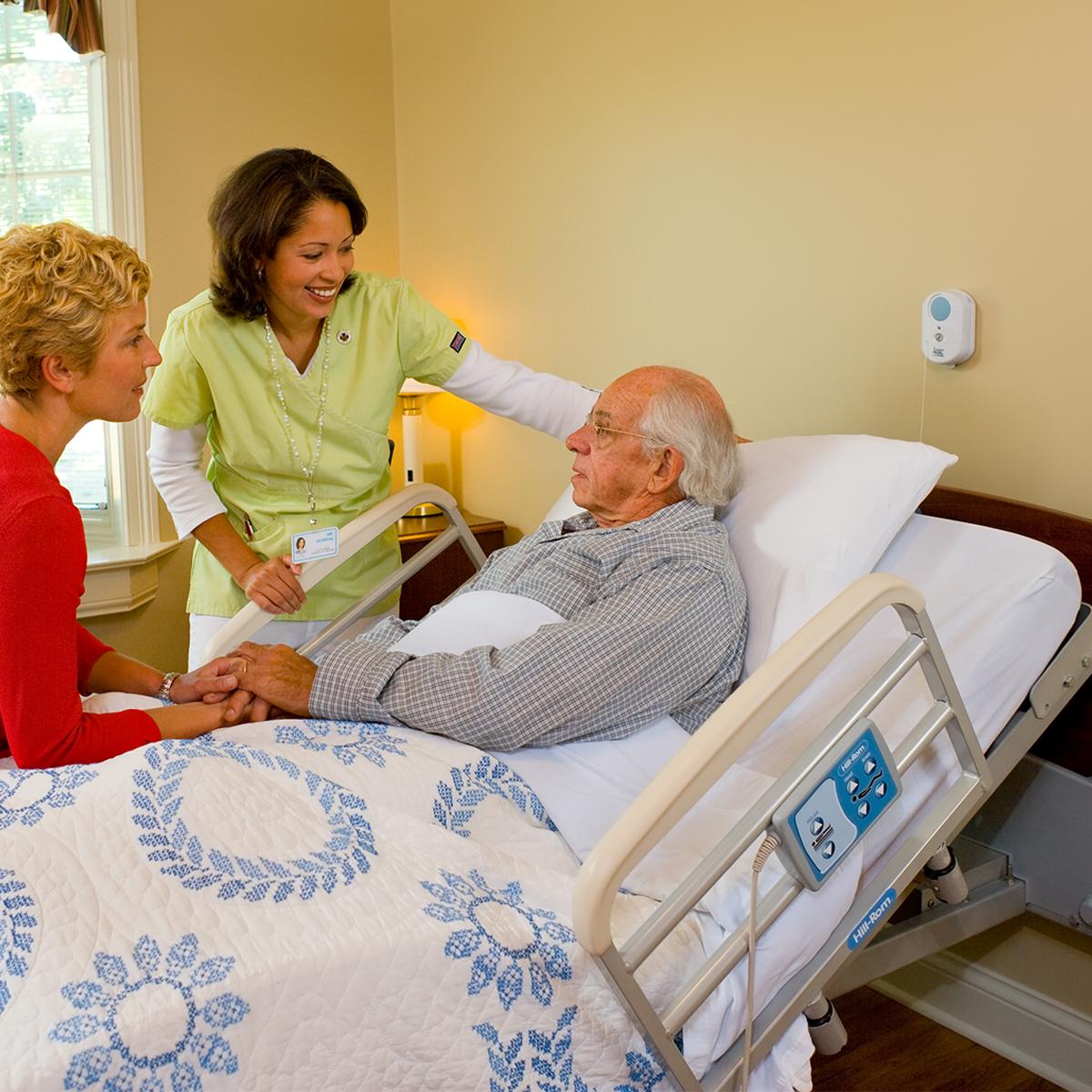 Older patient reclines in bed, talking with clinician and family member