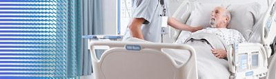 A clinician rests their hand on a patient's shoulder while the patient rests in a Hillrom hospital bed.