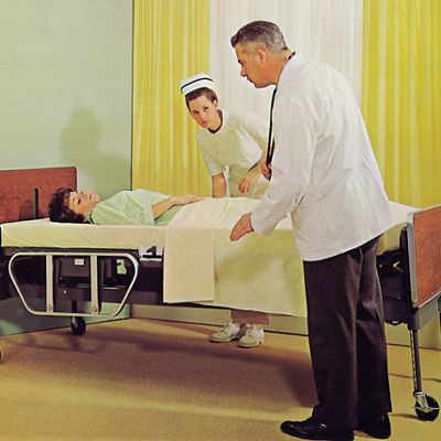 A doctor and nurse care for a patient lying in an early-model Hillrom bed