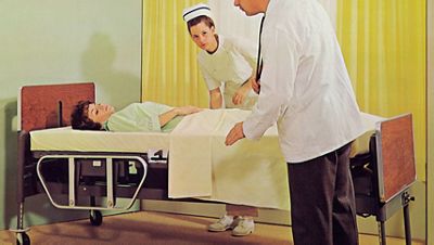 A doctor and nurse care for a patient lying in an early Hillrom bed