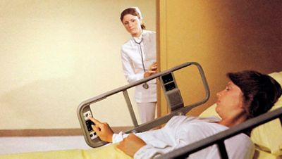 A nurse looks in to check on a patient pressing a nurse call control on an early Hillrom hospital bed siderail