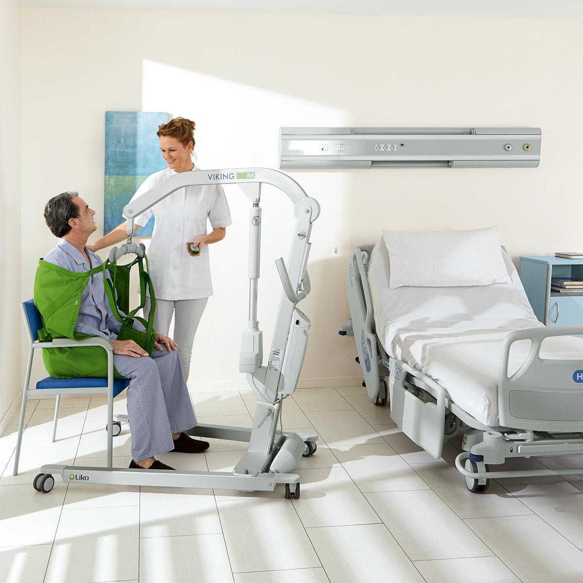 A patient sits in a seated sling attached to a patient lift while a clinician assists, with a Hillrom 900 bed in the room