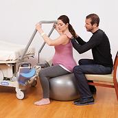 A pregnant woman sits on an exercise ball while holding a rail on an Affinity 4 Birthing Bed. A seated man has his hands on her shoulders