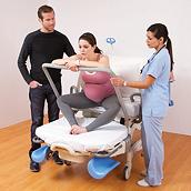 A pregnant woman sits in the Affinity 4 Birthing Bed and leans forward on a supporting rail, while a caretaker and man assist