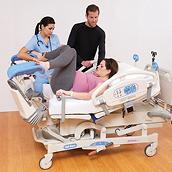 A pregnant woman lies in the Affinity 4 Birthing Bed with her legs raised on calf supports. A caretaker and man assist