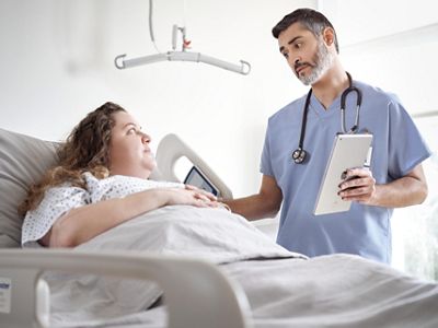 A clinician reviews information with a patient lying in bed, with an overhead lift in the background