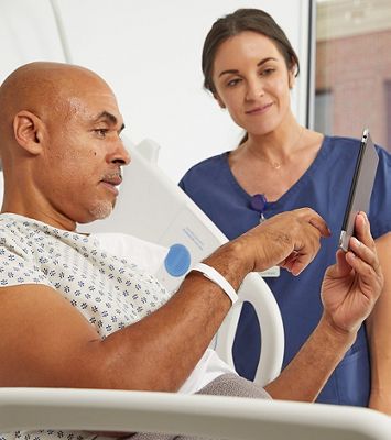 A patient in a hospital bed interacts with a tablet device while a caregiver watches.