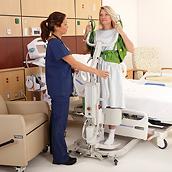 A clinician helps an older female patient stand from a hospital bed using a Sabina II mobile lift