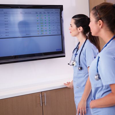 Two clinicians in blue scrubs review patient status data on a large screen in a hospital hallway