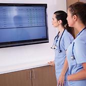 Two clinicians in blue scrubs review patient status data on a large screen in a hospital hallway