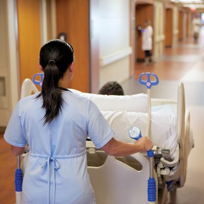 Una vista por encima del hombro de un cuidador que transporta una cama bariátrica Compella por un pasillo de hospital.