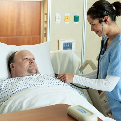 Bariatric patient lies in Excel Care ES Bariatric Hospital Bed while a clinician listens to his heart with a stethoscope