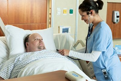 Bariatric patient lies in Excel Care ES Bariatric Hospital Bed while a clinician listens to his heart with a stethoscope