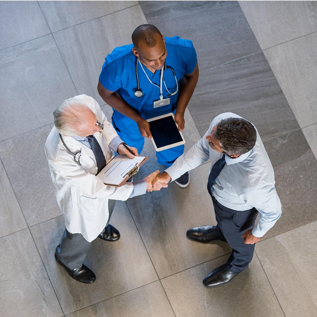 Clinician in blue scrubs with a tablet and physician in white lab coat, speaking to a man in business attire