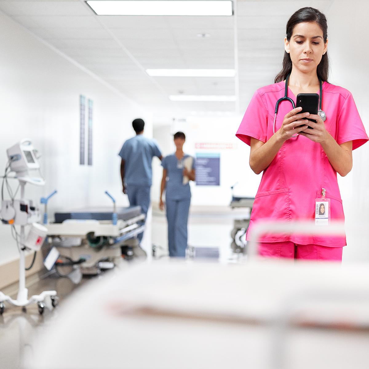 แพทย์หญิงตรวจสอบข้อความบนโทรศัพท์ของเธอขณะที่ยืนอยู่บริเวณทางเดินในโรงพยาบาล เธอสวมชุดสครับสีชมพู