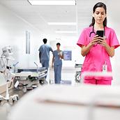 A clinician texts checks her phone while standing in a hospital corridor. She is wearing pink scrubs.