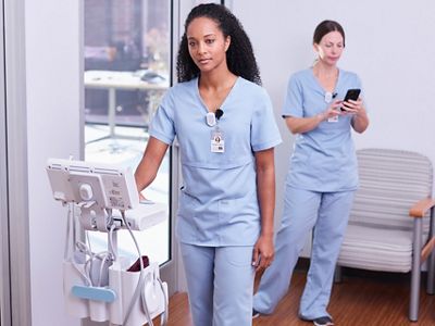 Two caregivers are in the hallway busy while wearing Hillrom Precision Locating badges and moving equipment tagged with the asset tag.