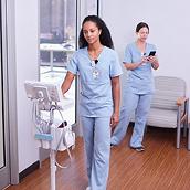 Two caregivers are in the hallway busy while wearing Hillrom Precision Locating badges and moving equipment tagged with the asset tag. 