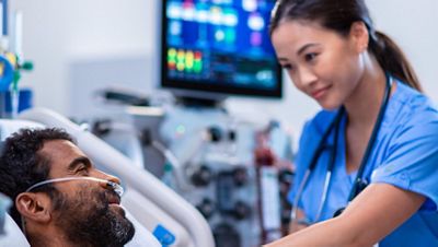 A gloved clinician checks on her patient in an ICU bed