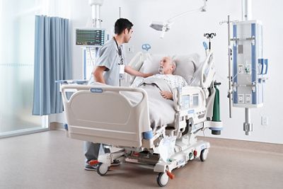 An older male patient lies in a Progressa bed with a clinician tending to him.