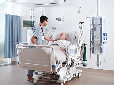 A caregiver rests his hand on the shoulder of a patient lying in a hospital bed