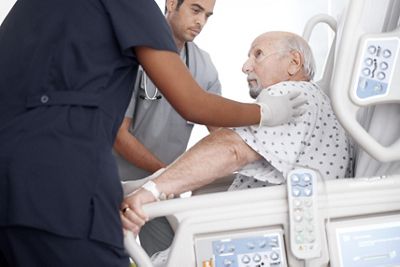 Two clinicians prepare to help a patient stand with the Progressa bed in FullChair position.