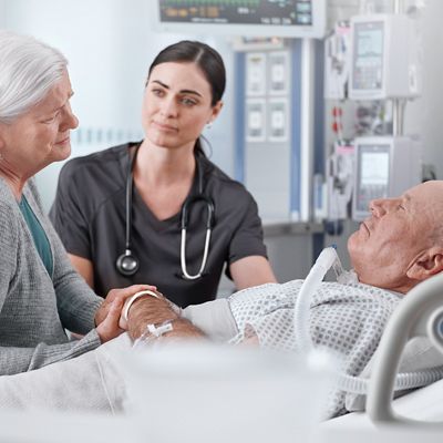 A woman holds the hand of a reclining male patient, caretaker in background