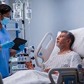 A masked clinician holding a tablet talks to a patient sitting up in a Progressa ICU bed.