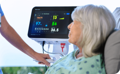older woman lying in hospital bed with nurse touching her shoulder with the connex 360 vital signs monitor next to the bedside