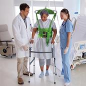 Ein Deckenlifter, der an einer grünen Weste befestigt ist, stützt eine Patientin mit einer Gehhilfe, wobei zwei Ärzte mithelfen