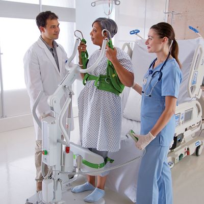 Two clinicians help an older female patient stand in a hospital room using a Sabina II mobile lift