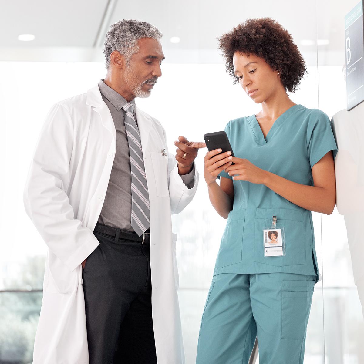 Two clinicians consult in a hospital hallway. One clinician holds her smartphone. Her male colleague points to its screen.