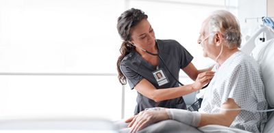 Un patient qui porte une canule nasale se tient assis et droit dans un lit de Hillrom pendant que sa clinicienne écoute son cœur avec un stéthoscope.