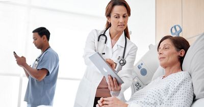 : A physician reviews information on a tablet with her patient while another clinician reads a message on his smartphone in the background.