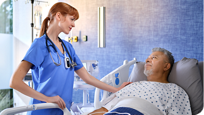 Clinician in blue scrubs stands beside a patient in a hospital bed, resting a hand on the patient’s shoulder in a clinical care setting.
