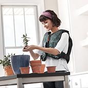 A smiling young woman with black hair, wearing a colorful Monarch® System vest and purple headband, pots plants in her living room.