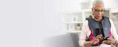 An older woman uses her smartphone while wearing the Monarch device with Zesty Pink vest in her family room