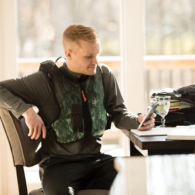 Smiling young man in gray activewear and the Adventure Green digital camouflage-pattern Monarch® System vest sits at table, using his smartphone.