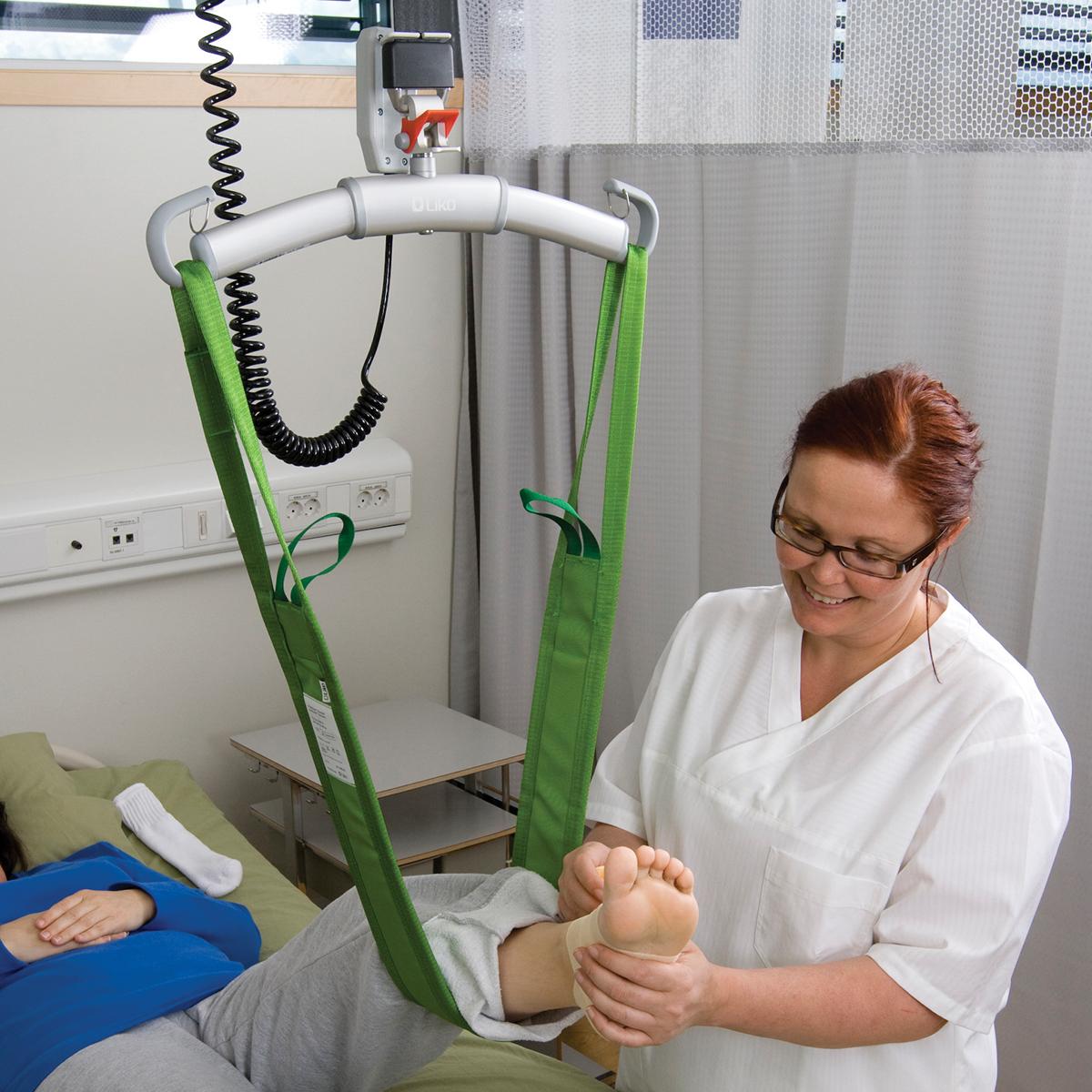 緑色の MultiStrap™ リフト補助具を使用して、リクライニング状態の患者さんの左足をストラップで支えている眼鏡をかけた女性医師