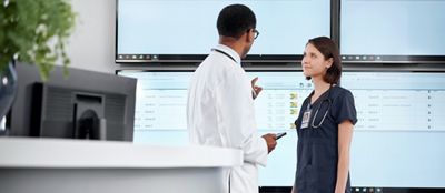 Two clinicians discuss patient statuses near a nurses’ station, with large monitors displaying Voalte Nurse Call.