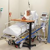A patient uses her phone while sitting upright in a Hillrom Procedural Stretcher. She rests her hands on an overbed table.