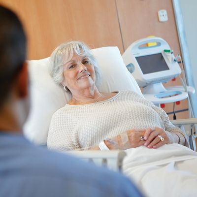 A clinician approaches a smiling patient in a Hillrom Procedural Stretcher. A Connex® Vital Signs Monitor is in the background.