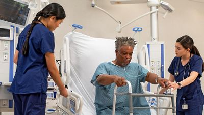 Two clinicians help a patient stand up from a Progressa+ ICU bed