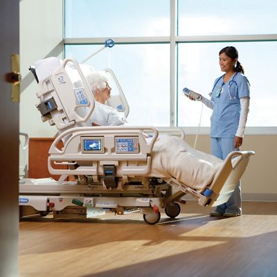 An elderly patient reclines in a Progressa bed in an upright sitting position while a caretaker operates the bed from a wired remote