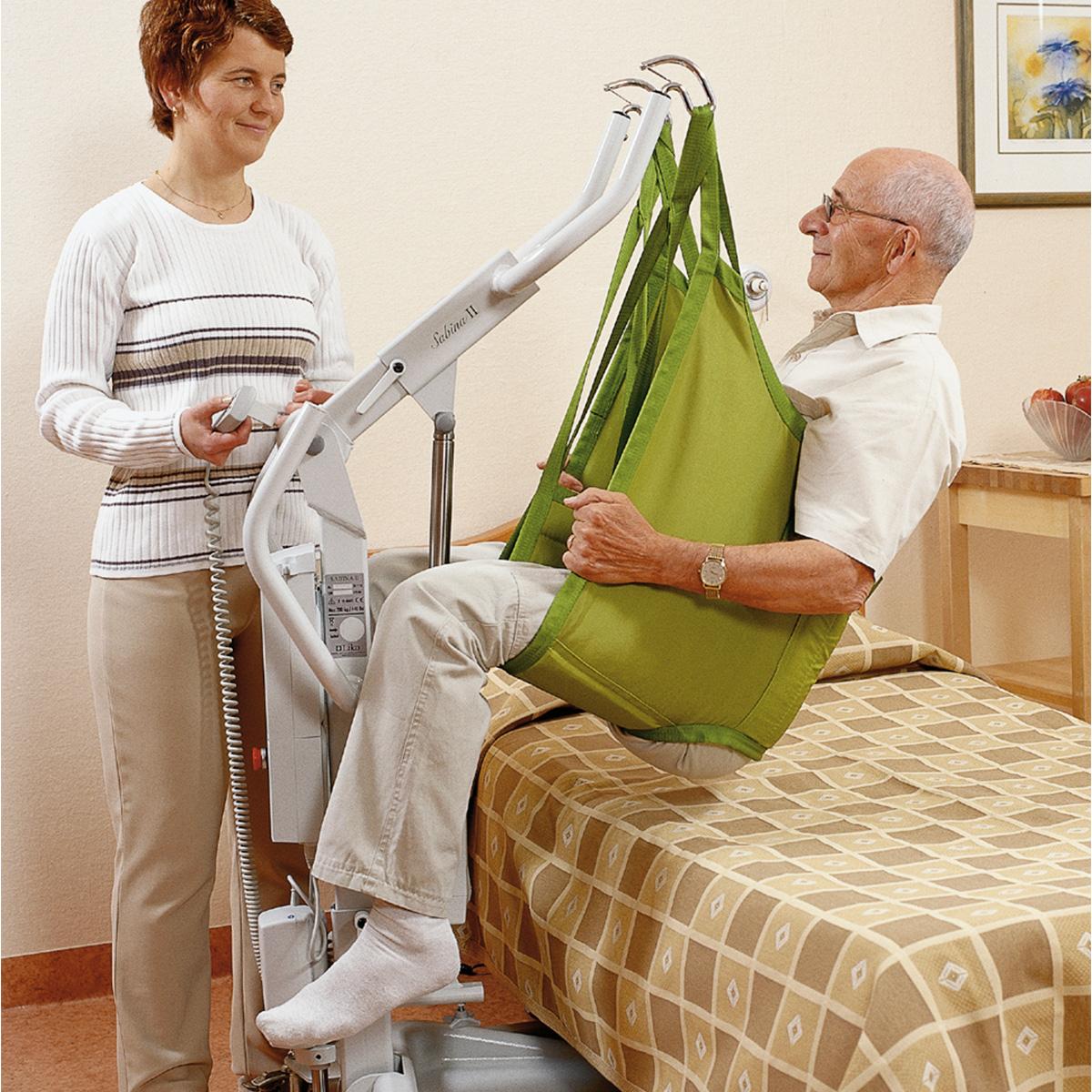 A family member helps an older male patient stand from a bed using a Sabina II mobile lift