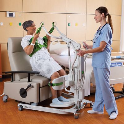 A clinician helps an older female patient stand from a chair using a Sabina II mobile lift