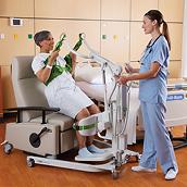 A clinician helps an older female patient stand from a chair using a Sabina II mobile lift