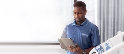 A Hillrom service technician consults a tablet from a hospital bedside