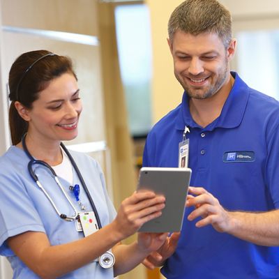 A care provider and a Hillrom support professional consult in a corridor. The clinician shows him data from her tablet device.