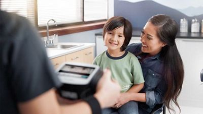 A child sits on a mother’s lap while the clinician sits across from them completing a vision screening with the Welch Allyn® Spot® Vision Screener.